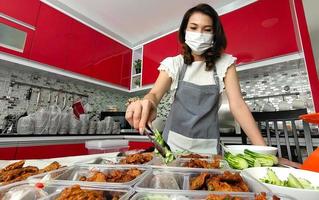 Asian housewife wearing hygiene mask preparing set of native Thai style food,  fried fish-paste patty, for sale ready to deliver for customers for extra income during coronavirus quarantine at home photo