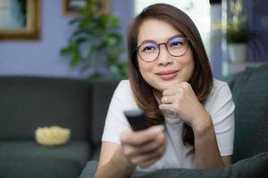 Cute Asian woman laying on sofa with relax and cozy gesture holding remote control aiming to tv, watching television and smile with fun and happy with popcorn blur in background. Hobby and relax idea photo