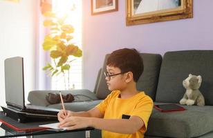 Young little boy wearing eyesglasses sitting near sofa in living room at home and do homework during class learning via laptop notebook computer photo