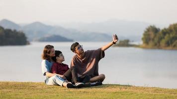 Asian family three members, mother and two young sons, sitting together beside huge lake with mountains and water in background. They using smartphone to take photos
