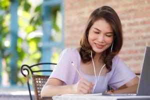 Cute beautiful middle-aged Asian woman sitting in shop and using a pencil to write on a paper notebook with a laptop computer and wearing earphones. Work from anywhere concept photo