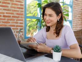Cute beautiful middle-aged Asian woman sitting in shop and using a pencil to write on a paper notebook with a laptop computer. Work from anywhere concept photo