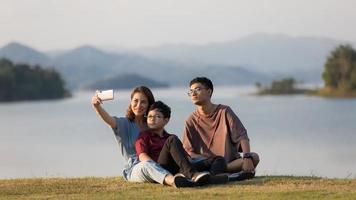 Asian family three members, mother and two young sons, sitting together beside huge lake with mountains and water in background. They using smartphone to take selfie photos