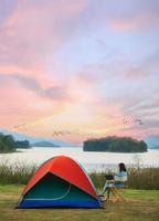 A sense of peace and serenity photo of female traveler sitting beside camping tent and using notebook laptop computer working from the lake side. There are group of birds flying in colorful sky