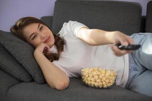 Beautiful and cute Asian woman reclining on sofa in living room and holding tv remote control with bowl of popcorn and sunlight shine from windows photo