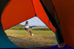 A woman sitting outside a camping tent and using laptop notebook computer working beside wide and big lake with clear blue sky and bids flying in natural scene wiith coffee cup. Taken from tent inside photo
