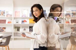 Portrait of Asain pretty cheerful female standing and reading book in library and leaning on glass wall with happy, focus and concentrate with reflection in clear mirror looking at camera photo