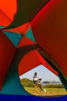 A woman sitting outside a camping tent and using laptop notebook computer working beside wide and big lake with clear blue sky and bids flying in natural scene wiith coffee cup. Taken from tent inside photo