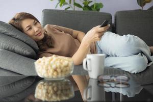 Asian woman holding remote control and looking at tv with happiness while laying on cozy sofa with popcorn and glass of drink at home living room photo