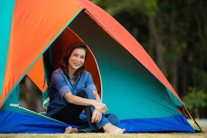 Beautiful Asian woman sitting and smiling in front of colorful tent with forest in background. Idea for tourist life in outddor adventure. photo