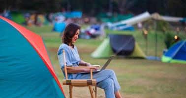 Female traveler sitting beside camping tent and using notebook laptop computer working with other tents blur in. background photo