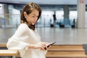 hermosa mujer madura asiática sentada en el interior y usando mensajes de texto de teléfonos inteligentes y enviando mientras espera a alguien. sonrisas femeninas atractivas y felices mientras mira la pantalla del teléfono móvil foto