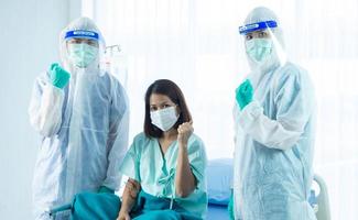 Asian woman patient feel happy because get well and recovered from COVID-19 or CORONA virus diease. Two medical staffs wear PPE suit standing beside patient bed and looking at the camera photo