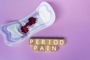 Period pain text on wooden block. Sanitary pads on purple background. Top view. photo