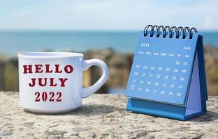 hola julio de 2022 escrito en una taza de café blanca con calendario azul foto