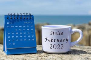 hola febrero de 2022 escrito en una taza de café blanca con calendario azul foto