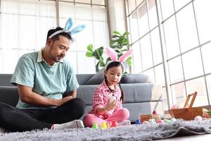 Father daughter preparing beautiful Easter eggs for decoration and celebration spring beginning, adorable pink little bunny girl kid and her dad with rabbit ears headband paint colorful eggs together photo