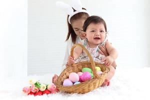 Happy smiling adorable six months baby girl playing with wicker basket of colorful Easter eggs in embrace of mother arms, mom with rabbit ears headband holding her sweet little daughter kid. photo