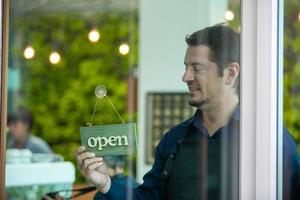 Portrait of smiling owner man standing at his cafe door with open signboard. Coffee owner standing in front of coffee shop to welcome customer and open the coffee shop in morning. photo