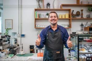 Portrait of smiling owner man standing at his cafe. Coffee owner standing with apron in coffee shop to welcome customer. photo