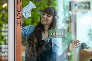 Portrait of smiling owner cleaning the door at her cafe with open signboard. Coffee owner in coffee shop to welcome customer and open the coffee shop in morning.