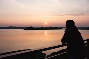 silhouette of hand of woman forming in heart shape around sun at sunset near lake photo