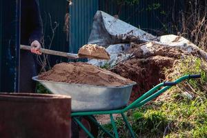 Man uses shovel and fills wheelbarrow with sand. Construction works. Rural life in countryside. photo