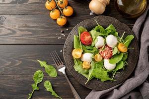 Fresh salad with arugula, cherry tomatoes, mozzarella cheese and hard cheese on dark wooden background. Top view photo