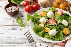 Fresh salad with arugula, cherry tomatoes, mozzarella cheese and hard cheese on white wooden background. Top view photo