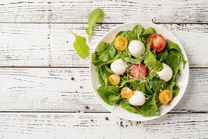 Fresh salad with arugula, cherry tomatoes, mozzarella cheese and hard cheese on white wooden background. Top view photo