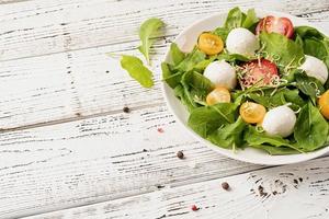 Fresh salad with arugula, cherry tomatoes, mozzarella cheese and hard cheese on white wooden background. Top view photo