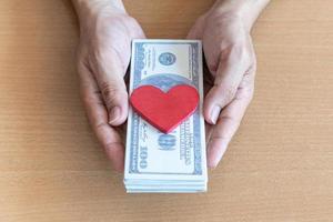manos de hombre sosteniendo billetes de 100 dólares y corazón rojo en una mesa de madera. da dinero de donación al concepto de donantes de corazón foto