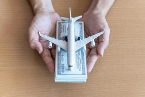 manos de hombre sosteniendo billetes de 100 dólares y avión en mesa de madera. concepto de viaje foto