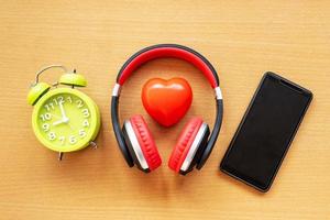 Headphones and alarm clock and smartphone and red heart on wooden desk. Musical concept photo