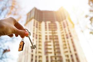 Hand with a key and a wooden key ring-house. Background of multi-apartment skyscraper. Building, moving to a new flat, mortgage, rent and purchase real estate. To open the door. Copy space photo
