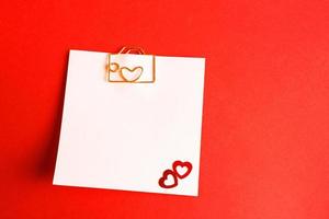 Square sheet for notes with a paper clip in shape of a letter and a heart. Decor of couples in love with hearts on a red background. Valentine's Day, message, greeting, declaration of love. Copy space photo