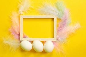 Easter white frame on a bright yellow background of chicken eggs and colored delicate feathers. Spring, religious holiday, Easter decoration, greeting, copy space, mock up photo