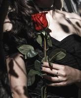 una chica con un vestido negro con una rosa roja. estilo gótico, mano femenina con uñas negras, símbolo de amor y muerte, tristeza, pérdida, estricta elegancia en negro