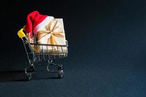 Box with a gift in Christmas packaging in a shopping cart on a black background. Black Friday, buying gifts for the new year. Space for text photo