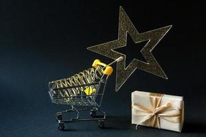 Shopping cart with a gold transparent star with sequins in the shape of a Christmas tree on a black background. Black Friday, buying gifts for the new year. Space for text photo