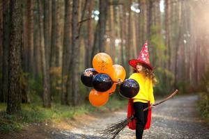 una niña con un disfraz de bruja y un sombrero en una escoba con globos naranjas y negros está jugando en el bosque de otoño, yendo a una fiesta de halloween foto
