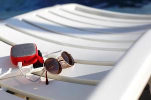 Red music portable speaker is charged from the power bank via usb on a deck chair near the pool. Concept is always in touch, travel gadgets, external battery for a smartphone. Place for text. Flatlay photo