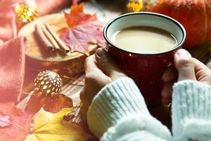 Red mug with hot cocoa on table with fallen maple leaves, round lamps, garlands, cinnamon sticks, warm blanket. Autumn atmosphere, warming coffee, women's hands in white sweater, cosiness and comfort photo