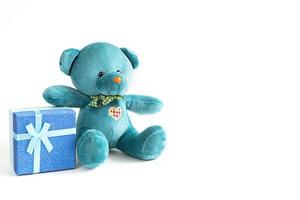 un oso de peluche turquesa suave con un corazón bordado sostiene una caja de regalo y un lazo sobre un fondo blanco. juguete para niños amor, un regalo de vacaciones, una declaración de amor, el día de san valentín. copie el espacio