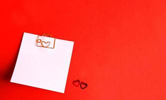 Square sheet for notes with a paper clip in shape of a letter and a heart. Decor of couples in love with hearts on a red background. Valentine's Day, message, greeting, declaration of love. Copy space photo