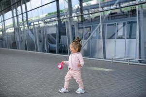 A little girl tries to walk on her own against the background of a modern building. In the hand of a girl holding a cup of spill.