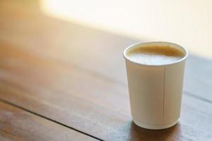 Closeup of disposable takeaway paper cup of hot coffee latte with milk foam on wooden table. photo