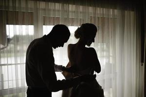 hermosa novia rubia con un lujoso vestido blanco preparándose para la boda. preparativos de la mañana. una mujer se pone un vestido. el novio ayuda a la novia. foto