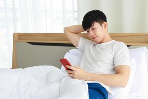 hombre asiático vestido con tela blanca usando un teléfono móvil para revisar las noticias matutinas y las redes sociales justo después de despertarse en su cama con espacio para copiar