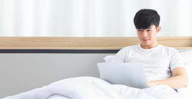 Young freelance Asian man working from home using laptop computer while sitting on the bed with copy space